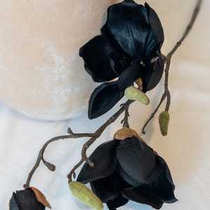 May include: A black artificial magnolia flower stem with two open blooms and a closed bud. The dark petals contrast with the light green buds and brown stems. The flower is placed on a white surface, with a beige vase in the background.
