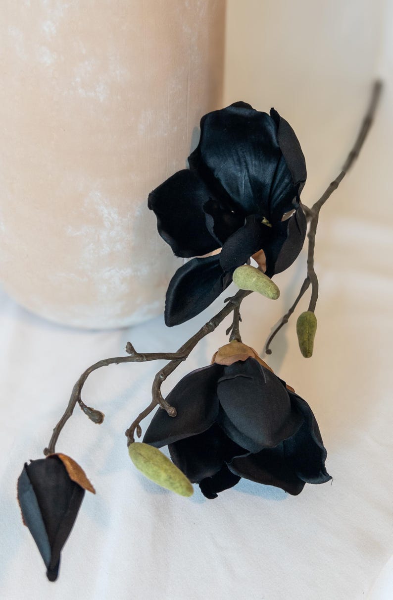 May include: A black artificial magnolia flower stem with two open blooms and a closed bud. The dark petals contrast with the light green buds and brown stems. The flower is placed on a white surface, with a beige vase in the background.