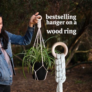 Puede incluir: Un colgador de plantas de macramé con una maceta negra que contiene una planta verde. El colgador está suspendido de un anillo de madera. El texto "bestselling hanger on a wood ring" es visible.
