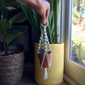 Puede incluir: Un macramé para colgar plantas con un anillo de madera, que sostiene una pequeña maceta de terracota con una planta verde. El macramé está hecho de cordón de algodón beige natural y tiene un borla en la parte inferior. El macramé está colgado de una mano frente a una ventana.