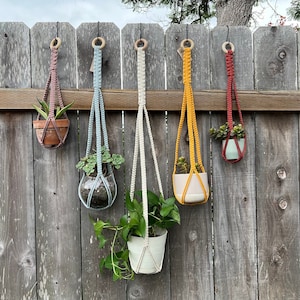 May include: Five macrame plant hangers in different colors hanging on a wooden fence. The hangers are made with thick rope and have a wooden ring at the top. Each hanger has a plant pot with a plant inside.