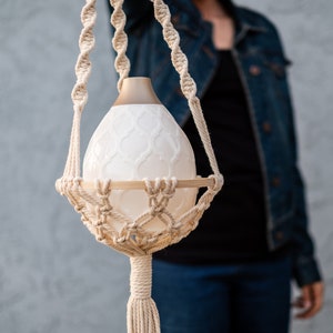 Op de afbeelding: Een witte keramische etherische olieverdamper met een gouden bovenkant, hangend aan een macramé plantenhanger.