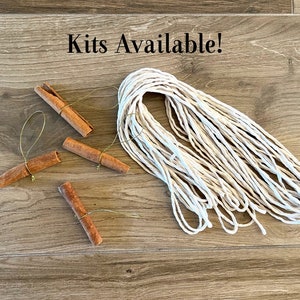 May include: A kit for making cinnamon stick ornaments includes four cinnamon sticks with gold string attached and a ball of white twine. The text "Kits Available!" is written on a wooden surface.