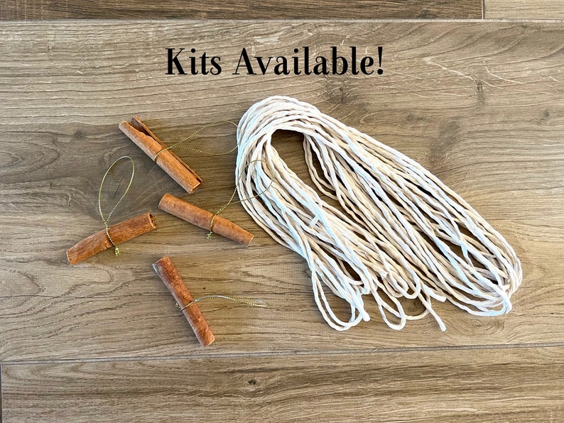 May include: A kit for making cinnamon stick ornaments includes four cinnamon sticks with gold string attached and a ball of white twine. The text "Kits Available!" is written on a wooden surface.