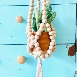 May include: A hanging planter with a cactus. The terracotta pot is suspended by a beaded wooden hanger. The hanger has a white tassel at the bottom. The background is a light blue dresser.