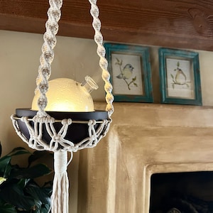 Może przedstawiać: Biały wiszący dyfuzor z makramą, z przezroczystym szklanym dyfuzorem i drewnianą podstawą. Dyfuzor wisi na białym sznurku z makramą i jest podświetlony. Dyfuzor znajduje się przed kominkiem z brązowym kamiennym obramowaniem.