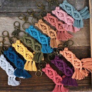 May include: A collection of colorful macrame keychains with tassels. Each keychain features a unique color combination and a braided design. The keychains are attached to gold-toned key rings.