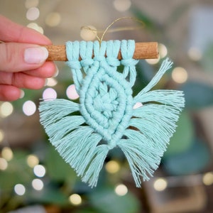 May include: A light blue macrame ornament with a cinnamon stick at the top. The ornament is in the shape of a butterfly with fringe details.