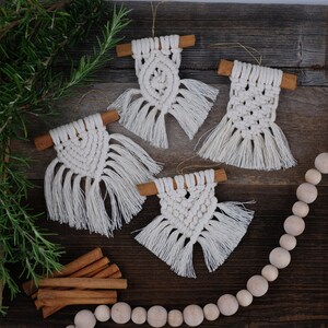 May include: Four white macrame ornaments with cinnamon stick hangers. The ornaments are hanging on a wooden surface with a strand of wooden beads and a sprig of rosemary.