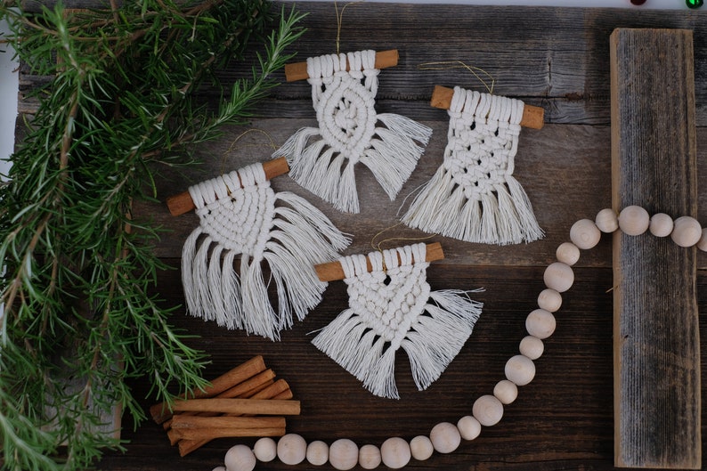 May include: Four white macrame ornaments with cinnamon stick hangers. The ornaments are hanging on a wooden surface with a strand of wooden beads and a sprig of rosemary.
