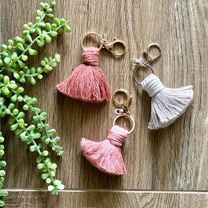 May include: Three tassel keychains with gold and silver key rings. The tassels are in shades of pink and beige.