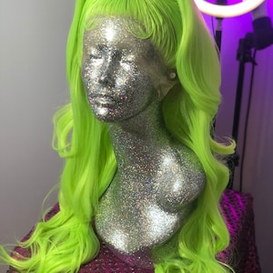 May include: A bright green wig with long, wavy hair. The wig is styled with a high ponytail and is displayed on a mannequin head covered in silver glitter.