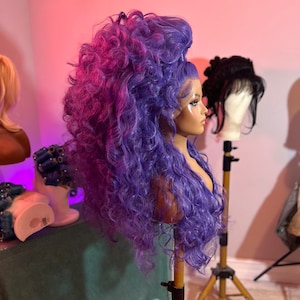 May include: A purple and pink curly wig on a mannequin head. The wig is styled in a high bouffant with large curls.