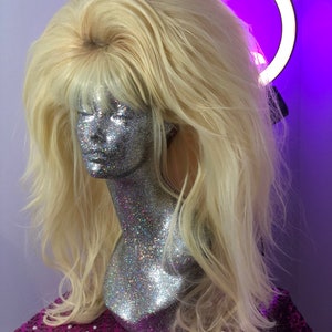 May include: A long, blonde wig with a layered, wavy style. The wig is styled with a side part and a fringe. The wig is displayed on a mannequin with a silver glitter finish.