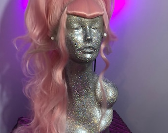 May include: A light pink wig styled with a high braided ponytail and long, wavy curls. The wig is displayed on a glittery mannequin head. The background is a light purple wall and a sequined surface.