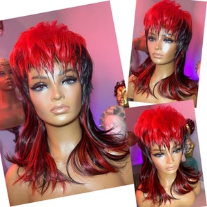 May include: A red and black mullet wig on a mannequin head. The wig features a layered style with a vibrant red top and black roots and sides. The mannequin has a neutral skin tone and is set against a soft pink background.