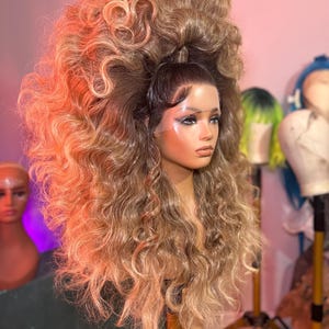 Może przedstawiać: Głowa manekina z dużą, obszerną peruką. Peruka ma mieszankę jasnobrązowych i blond odcieni z długimi, kręconymi pasmami. Włosy są ułożone wysoko, tworząc efektowny wygląd. Inne peruki są widoczne w tle.