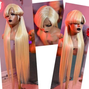 May include: A blonde wig with a blunt bang and long, straight hair. The wig is styled on a mannequin head with dark makeup. The hair is a light, golden color, and the style is sleek and modern. The wig appears to be made of high-quality synthetic or human hair.