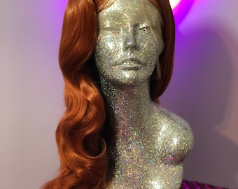 May include: A long, wavy, auburn-colored wig with a center part. The wig is styled in a vintage, glamorous look with soft, flowing curls. The wig is displayed on a silver glitter mannequin head.