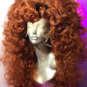 May include: A long, curly, auburn-colored wig with a large volume of hair. The wig is styled with large, loose curls and is worn on a mannequin head.