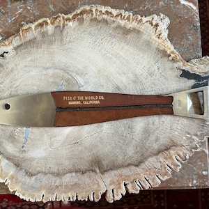 May include: A silver bottle opener with a brown leather handle. The handle has the text "FISH OF THE WORLD CO. BANNING, CALIFORNIA" embossed in gold.
