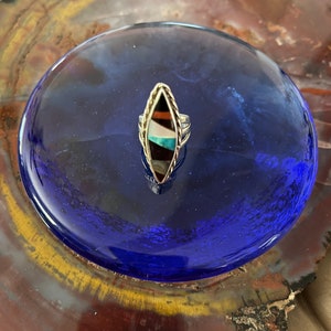 May include: A silver ring with a multi-colored inlay in the shape of a marquise. The inlay features black, brown, white, turquoise, and gold colors.