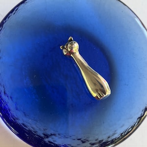 May include: A silver cat figurine on a blue glass dish. The cat is facing the right side of the image and has a long tail.