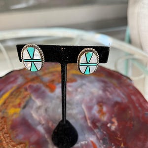 May include: Oval-shaped stud earrings with a silver rope-like border. The earrings feature a white background with a turquoise and black geometric design. The earrings are displayed on a black stand.
