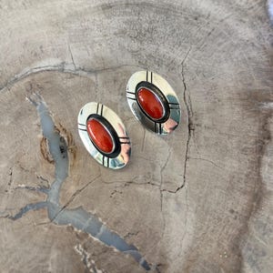 May include: A pair of oval-shaped earrings with a silver-tone metal frame. Each earring features a vibrant, oval-shaped, red stone in the center. The silver frame has a geometric design with vertical lines. The earrings are displayed on a wooden surface.