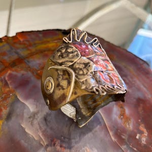 May include: A copper and brass fish sculpture with a textured surface and a red, orange, and yellow patina. The sculpture is made of hammered metal and has a unique, organic shape.