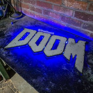 Może przedstawiać: Metalowy znak z napisem "DOOM" w postarzonej, białej czcionce, podświetlony niebieskimi światłami LED. Znak umieszczony jest na ciemnej powierzchni, z ceglaną ścianą w tle. Widoczny jest tekst "@GhostShopUS".