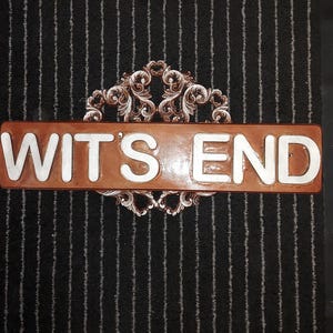 May include: A brown rectangular sign with the words "WITS END" in white lettering. The sign is framed by an ornate, decorative border. The background is a black rug with vertical white stripes.