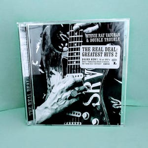 May include: A CD case featuring Stevie Ray Vaughan & Double Trouble's "The Real Deal: Greatest Hits 2." The album cover is black and white, showing a close-up of a hand playing a guitar. The text "BRAND NEW!!" is visible.