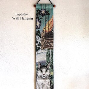 May include: A tapestry wall hanging with a detailed illustration. The top features a teal background with purple flowers and a decorative urn. The bottom shows a cat with black and white fur. The text "Tapestry Wall Hanging" is on the left.