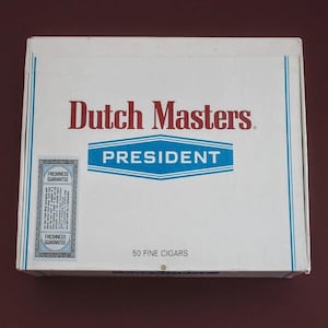 May include: A white box of Dutch Masters President cigars. The box has blue and red text, with the words "Dutch Masters" in red and "President" in blue. The box also has a "Freshness Guarantee" sticker and the text "50 Fine Cigars".
