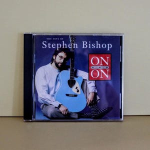 May include: CD case featuring the album "The Hits of Stephen Bishop." The cover art shows a man with glasses and a beard holding a light blue guitar. The album title and "ON AND ON" are displayed in white and red text.