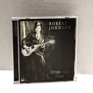 May include: A CD case featuring a black and white image of Robert Johnson playing a guitar. The text on the cover reads "ROBERT JOHNSON" and "Cross road Blues". The image is in a clear plastic case.
