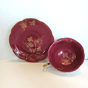 May include: A burgundy teacup and saucer set with a gold vine and grape design.