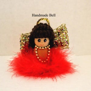 May include: Handmade bell ornament featuring a wooden doll with a brown afro, gold beaded necklace, and red feather skirt. The doll has a painted face and is adorned with gold glitter wings and a gold halo.