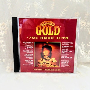 May include: A CD case with a red cover featuring the text "70 Ounces of GOLD" and "'70s ROCK HITS". The cover art includes a graphic of gold coins and a list of song titles by various artists. The CD case has a black spine.
