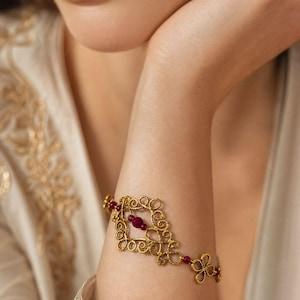 Puede incluir: Una pulsera de tono dorado que presenta un diseño intrincado de metal estilo filigrana con una superficie texturizada. La pulsera está acentuada con pequeñas cuentas redondas de color rojo intenso integradas en los patrones metálicos ornamentados.