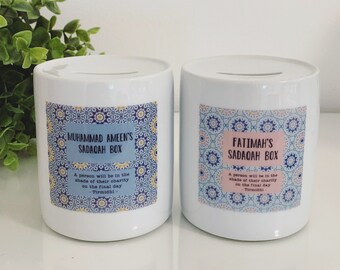 Puede incluir: Dos cajas Sadaqah de cerámica blanca con una ranura en la parte superior. Cada caja tiene una etiqueta decorativa con texto. Una dice "Sadaqah Box de Muhammad Ameen" y la otra "Sadaqah Box de Fatimah". Ambas tienen una cita de Tirmidhi.