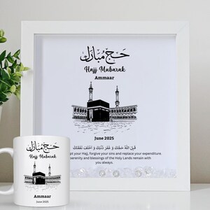 Puede incluir: Una impresión enmarcada en blanco y una taza blanca con una ilustración en blanco y negro de la Kaaba en La Meca. La impresión y la taza tienen el texto árabe "Hajj Mubarak", el nombre "Ammaar" y la fecha "Junio 2025".