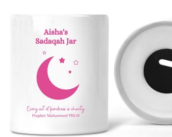 May include: White ceramic charity jar with a pink crescent moon and stars design. The jar has a slot on the top for inserting coins. The text on the jar reads "Aisha's Sadaqah Jar" and "Every act of kindness is charity. Prophet Muhammad PBUH".