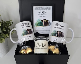 May include: A black gift box with a white mug, a candle, and chocolate hearts. The mug and candle have Arabic calligraphy that says "Umrah Mubarak".