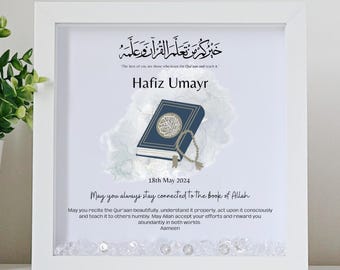 May include: A white framed print with a blue and white illustration of a book and prayer beads. The text reads "The best of you are those who learn the Qur'an and teach it."  "Hafiz Umayr"  "18th May 2024"  "May you always stay connected to the book of Allah"  "May you recite the Qur'aan beautifully, understand it properly, act upon it consciously and teach it to others humbly. May Allah accept your efforts and reward you abundantly in both worlds"  "Aameen"