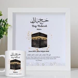 Könnte beinhalten: Ein weißer gerahmter Druck und eine weiße Tasse mit der Kaaba, einem schwarzen kubusförmigen Gebäude, mit goldenen Akzenten. Der Druck und die Tasse enthalten arabische Kalligraphie und den englischen Text "Hajj Mubarak" und "Juni 2025 | Dhul Hijjah 1446".