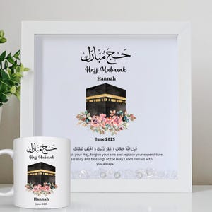 May include: A white framed print and a white mug with a Hajj Mubarak design. The design features a black and gold Kaaba illustration surrounded by flowers, with the name "Hannah" and "June 2025" below. The print has Arabic text.