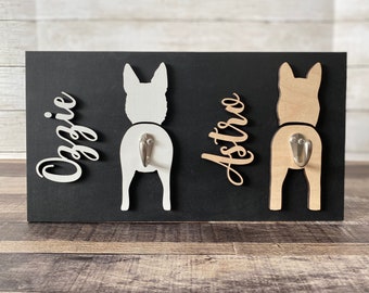 May include: A black wooden sign with three dog silhouettes, each with a metal hook. The silhouettes are painted white and light brown. The names "Oggie" and "Astro" are written on the sign.