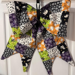 May include: A large, quilted Halloween bow featuring a patchwork design. The bow incorporates various fabrics with spooky patterns, including ghosts, bats, and spiderwebs in black, white, orange, green, and purple. The bow is a decorative item, likely for door or wall decor.